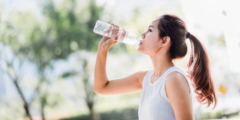 Woman in white tank top drinking water from a clear bottle outdoors.